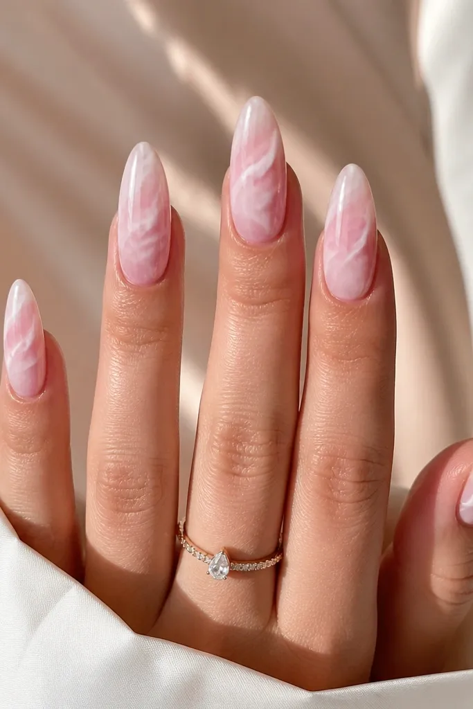 Almond nails painted milky pink with soft rose quartz swirls. The veining is creamy white and pale blush, flowing diagonally across each nail. The finish is glossy with a glass-like shine.