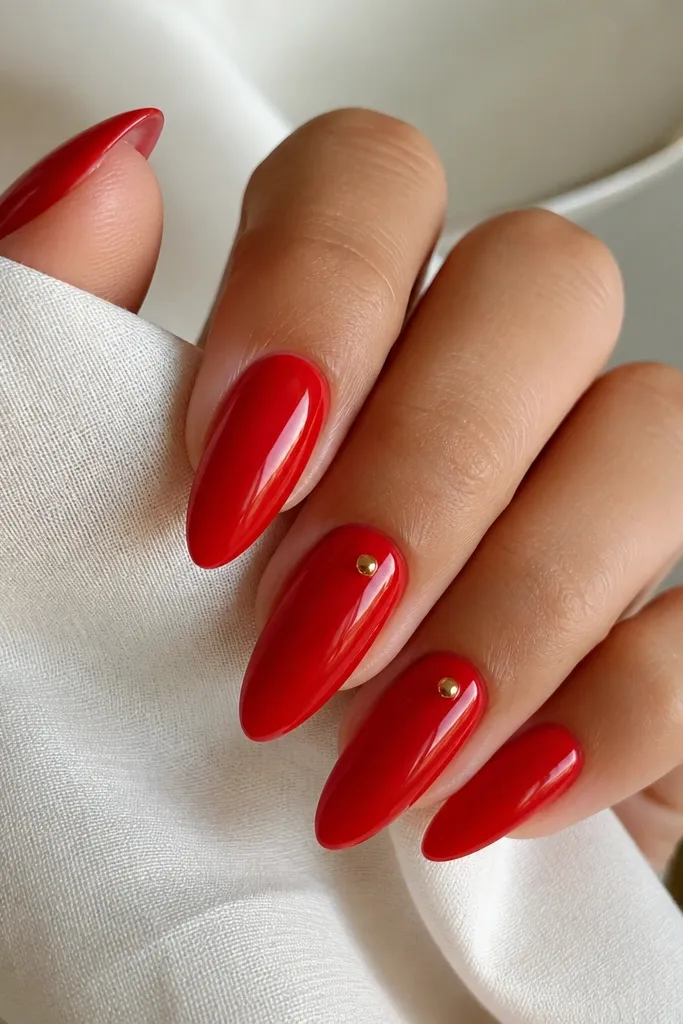 Long almond nails painted glossy red with a few tiny gold studs placed near the cuticle on two accent nails.