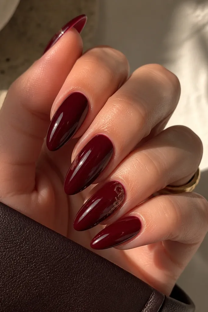 Long almond nails in deep black cherry red with mirror gloss, plus a tiny swirl design on one or two accent nails near the cuticle.