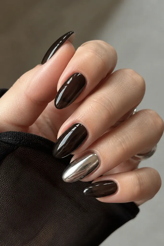 Short-to-medium almond nails in deep black jelly finish, with the ring finger covered in mirror silver chrome. The black nails look translucent at the edges, and the chrome ring finger reflects bright light like metal.