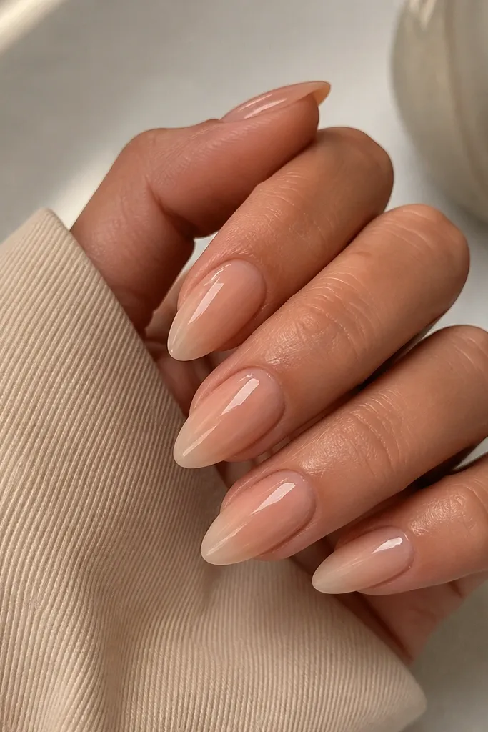Almond nails in a warm nude tone that matches the skin's warmth. The finish is glossy and evenly colored, with a neat cuticle boundary. The free edge looks slightly lighter than the base, like a soft gradient.
