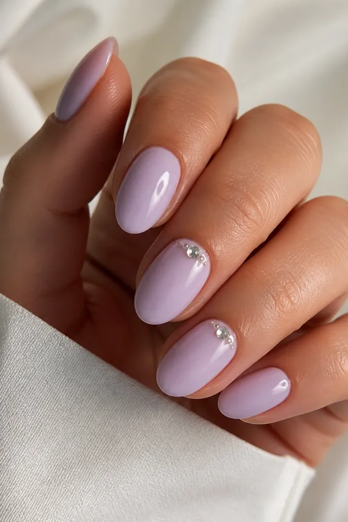 Short almond nails painted lavender milk. On two nails there are tiny clear rhinestones placed in a small cluster near the cuticle center, with one or two micro dots of white gel. The nails are glossy and clean.