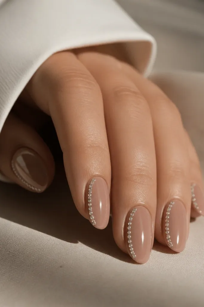 Short almond nails in nude brown. Along only the left side of each nail there's a thin row of tiny clear micro rhinestones from cuticle to almost the tip. The right side is plain nude, and everything is glossy.