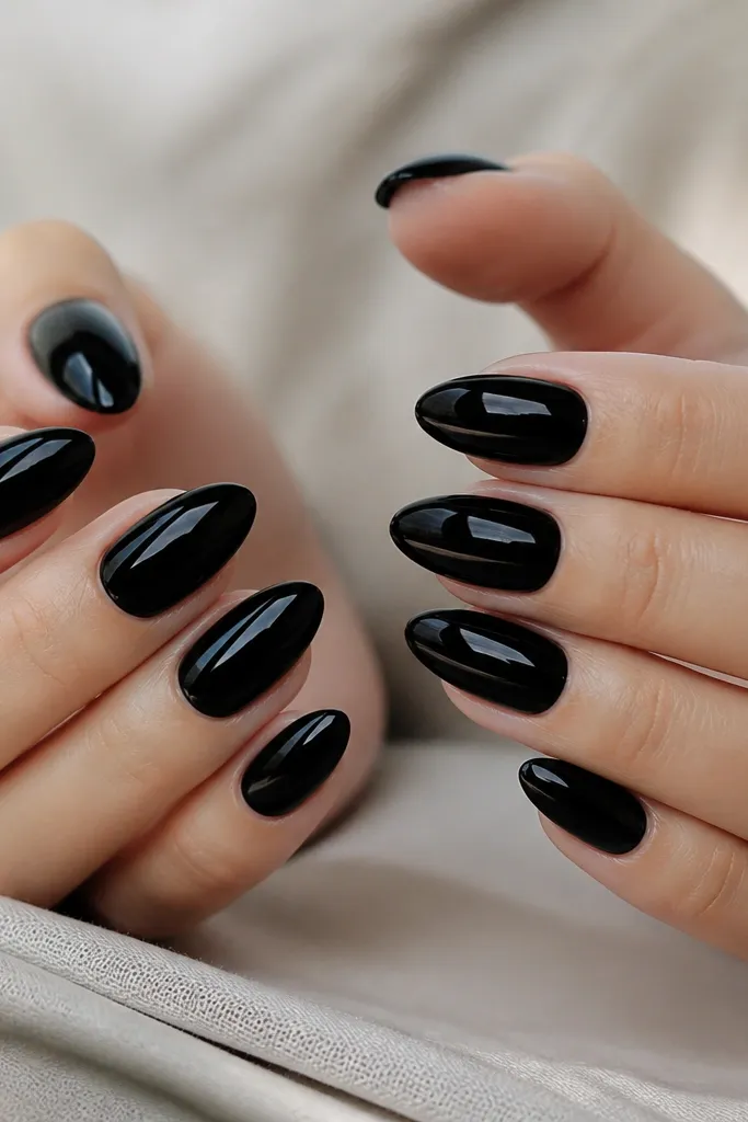 Close-up of almond-shaped nails painted solid black with a mirror-like shine. The cuticles are clean and the surface looks smooth, like glass. No gems, just a perfect glossy finish on all ten nails.