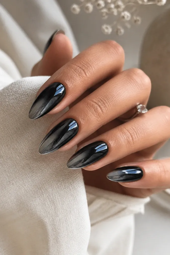 Almond nails coated in black chrome. There's a subtle silver fade toward the tips, like a smoky gradient under the chrome finish. High shine catches light across the surface.