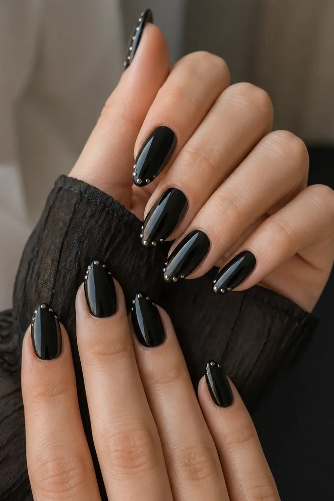 Almond nails in solid glossy black. Each nail has 2-3 tiny silver studs clustered along the outer tip edge, like a subtle sparkle halo. Cuticles are clean and the surface is smooth.