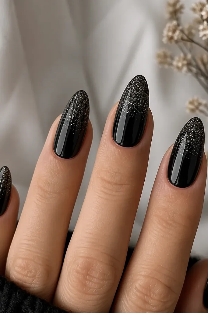 Almond nails with a black base that fades into fine black glitter near the tips. The glitter is concentrated at the ends and disappears smoothly toward the cuticle. Glossy topcoat makes it sparkle.