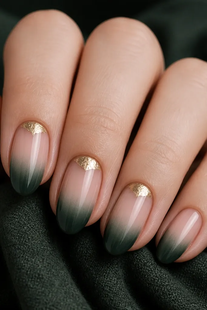 Close-up of nails over a dark green scarf. The nails are medium-short almond. The base is sheer nude, and the tips are painted deep evergreen with a smooth gradient edge. On three nails, gold foil half-moons sit at the cuticle line, partially covering the nude base.