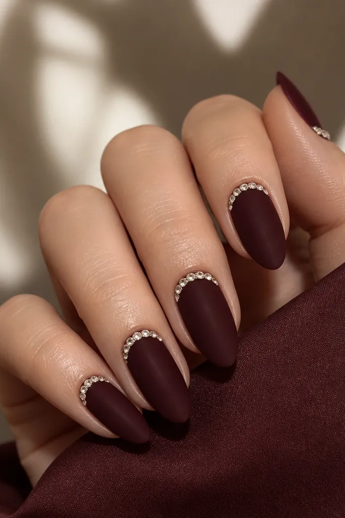 Medium almond nails in deep wine with a subtle velvet-matte finish; on each nail there's a silver half-moon near the cuticle made of small rhinestones. The rest of the nail is smooth and dark, with no glitter.