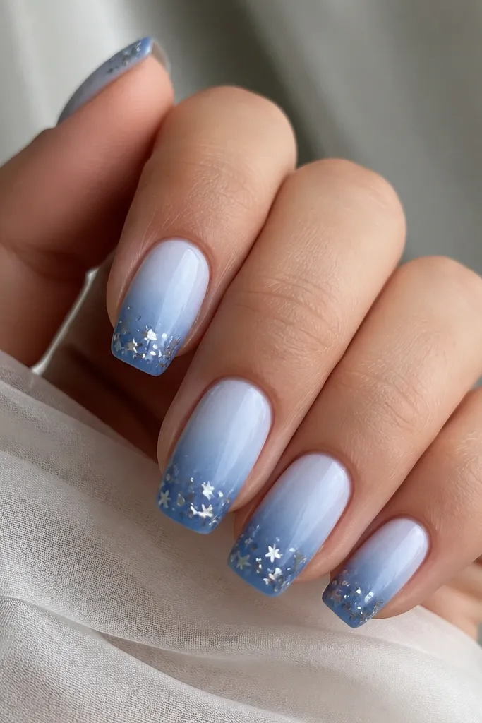 Medium squoval nails with a glacier blue ombré fading from pale icy blue near the cuticle to deeper blue at the tip; each nail has a few small star-shaped foil pieces clustered at the free edge.