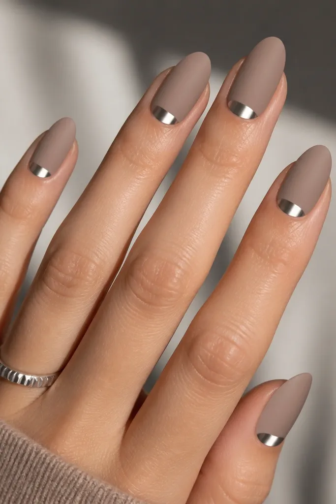 Medium almond nails in soft taupe; each nail has a clean icy silver half-moon at the cuticle area. The chrome is reflective and smooth, and the taupe base stays matte or satin.