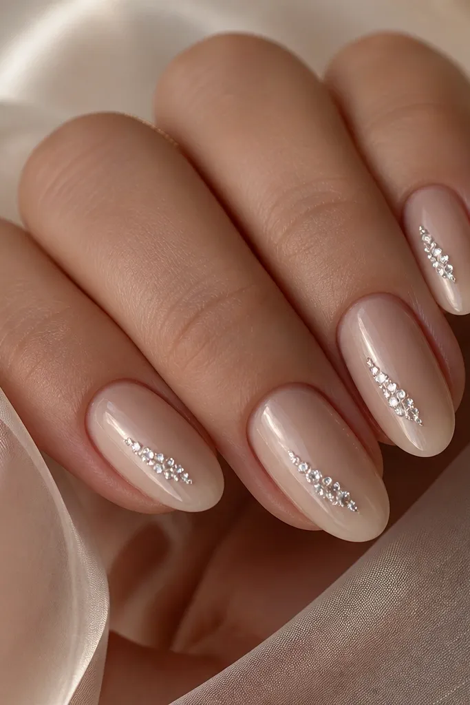 Short almond nails painted champagne nude with a high-gloss finish; each nail has a small crystal-clear rhinestone cluster placed diagonally from mid-nail toward the tip. The rhinestones catch light like glass.
