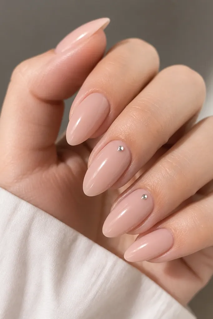 A close-up of nude-milky pink almond nails with a single tiny silver dot centered right at the cuticle on two accent nails; glossy finish, smooth and clean edges.