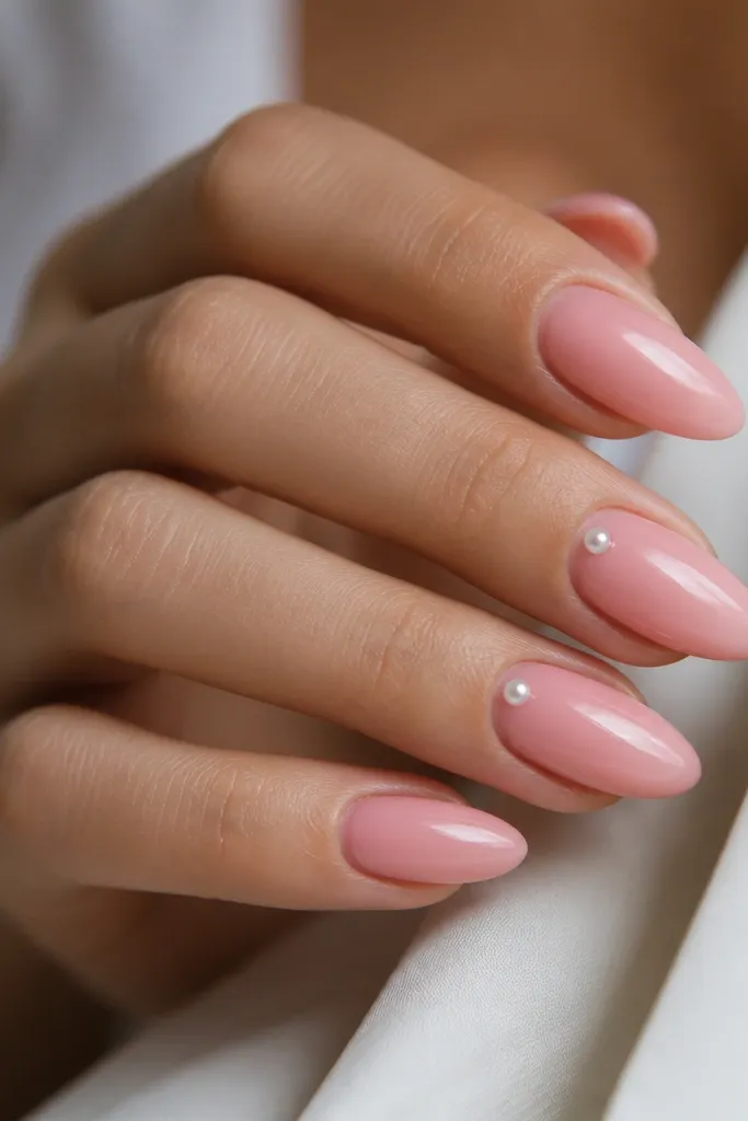 Glossy pink almond nails with one tiny pearl bead placed right on the cuticle edge on two nails; the rest is plain pink with crisp cuticles.