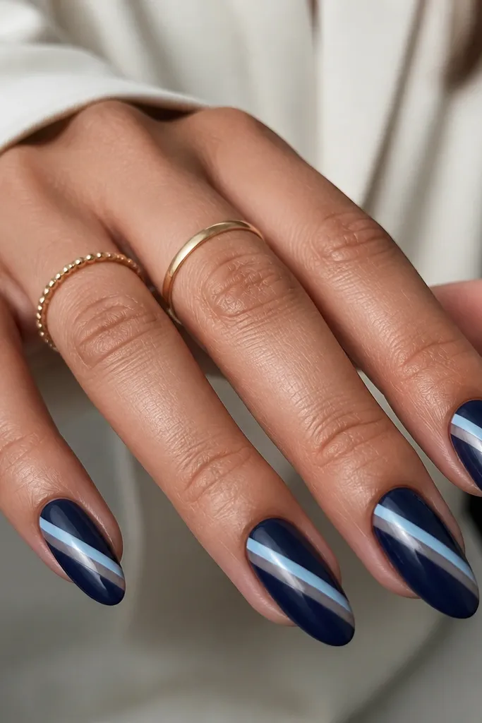 Almond nails painted deep navy with a diagonal band of negative space from lower left to upper right. Inside the diagonal band, there is a single icy-blue stripe running the length, with crisp edges.