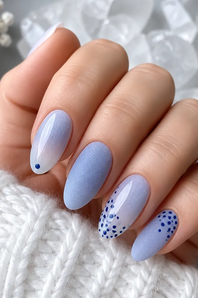 Five nails in a gradient mix of frosted periwinkle, powder blue, and clear icy base; two nails have tiny navy dot clusters like falling snow, one nail has a single navy dot at the tip, and the accent nails show a matte frosted finish with a glossy ice topcoat on the other nails.