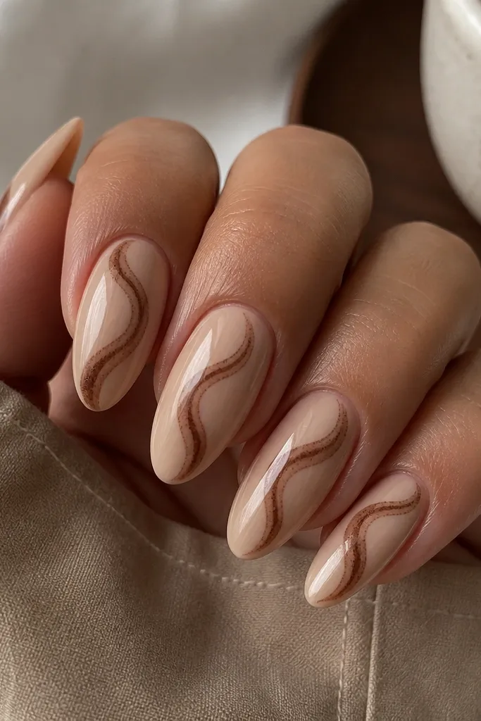 Glossy almond nails in a warm beige nude base. Each nail has a thin mocha-brown swirl that starts near the cuticle and curls toward the tip, with a subtle glazed shine and tiny micro-speckle texture in the swirl.