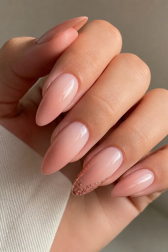 Long nude almond nails in a smooth nude gradient from sheer cuticle to slightly deeper nude tips. The ring finger has rose-gold foil fragments placed along the free edge like a thin border; other nails are plain glossy nude.