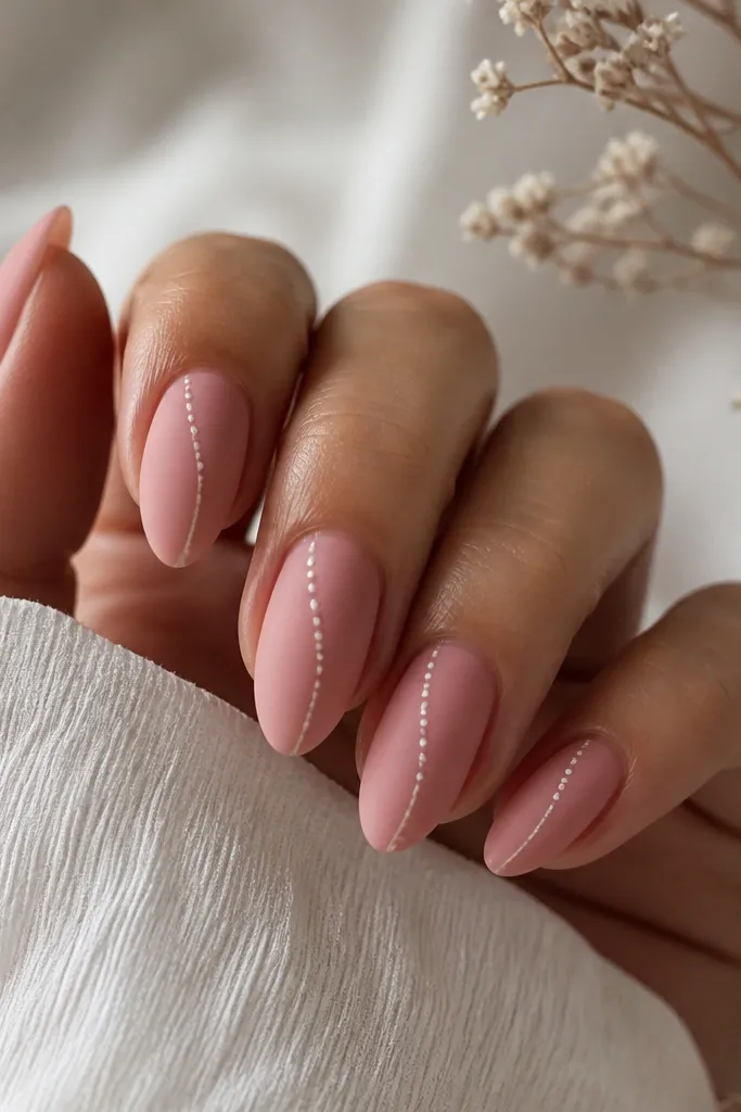 Dusty rose pink almond nails with a soft matte base finish. Each nail has a diagonal line of tiny white dots starting near one side of the cuticle and moving toward the center of the tip. The dots are small and evenly spaced.