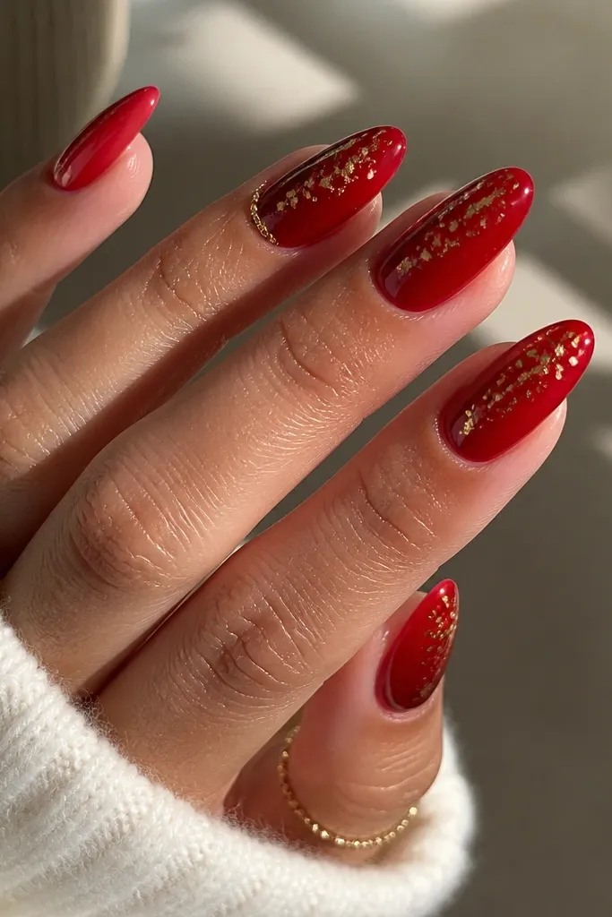 Almond nails in glossy glazed red; scattered gold foil flecks sit mostly near the upper half of each nail.