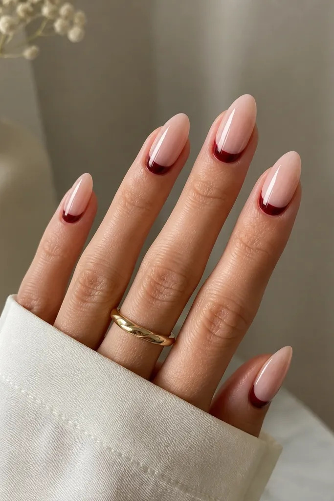 Almond nails with a sheer nude base and a wine-red half-moon at the cuticle. The half-moon is crisp, slightly rounded, and the rest of the nail is glossy and clean.