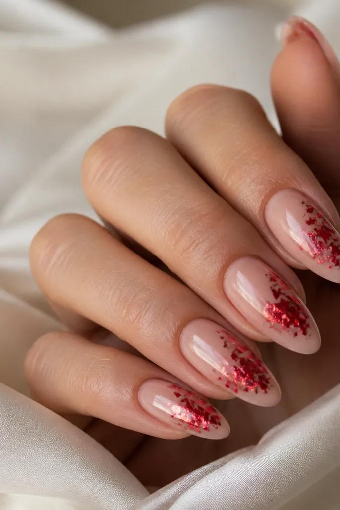 Almond nails in glossy sheer nude with a red foil flake cluster on one side of each nail, near the middle to tip. The foil catches light with sharp metallic reflections.