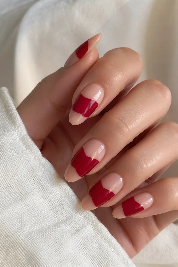 Almond nails with a nude base and a cherry-red reverse French band across the center, leaving a small nude triangle pointing toward the cuticle. The band is crisp and glossy.
