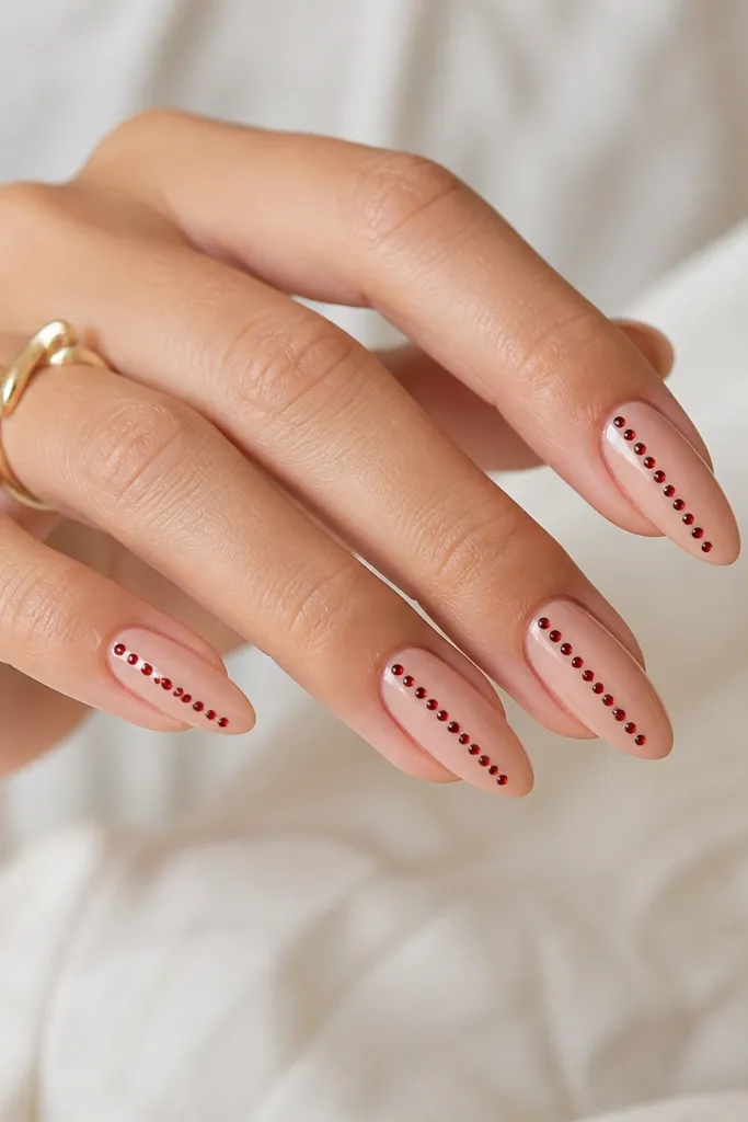 Almond nails with glossy nude base and a single line of tiny red rhinestones running vertically along one sidewall from mid-nail toward the tip. Each stone is spaced evenly.