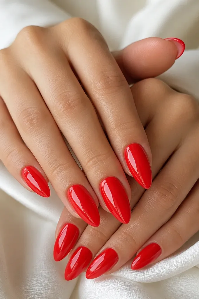 Ten almond nails painted a bright tomato red with a high-gloss, mirror-like finish. The nails are medium length, with crisp edges and a smooth tapered tip; no extra art.
