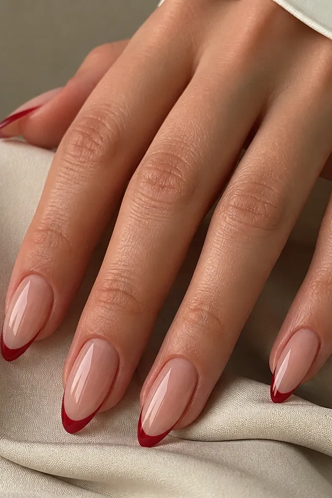 Almond nails with a sheer nude base. The tips have a thin cherry red French line shaped in a crisp curve, with the rest of the nail clean and glossy.