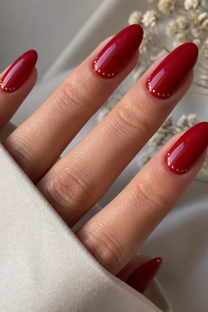Almond nails in glossy red with tiny gold micro dots clustered near the cuticle on each nail. The dots are spaced evenly and stay small.
