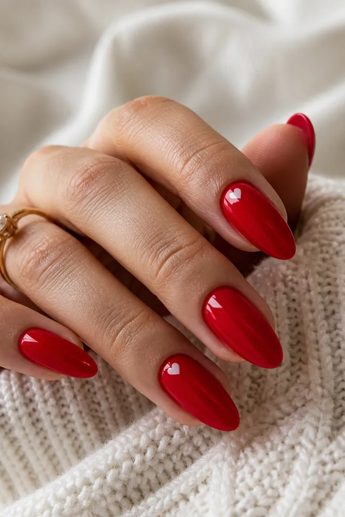Almond nails in glossy red with one or two nails featuring a tiny white heart near the cuticle. The hearts are small and centered, with clean edges.