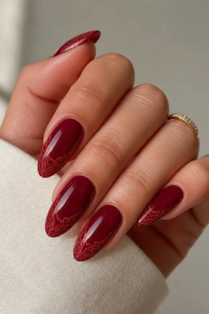 Almond nails painted deep burgundy red with thin lighter red lace-like linework. The pattern is delicate around the edges and curls toward the tip.