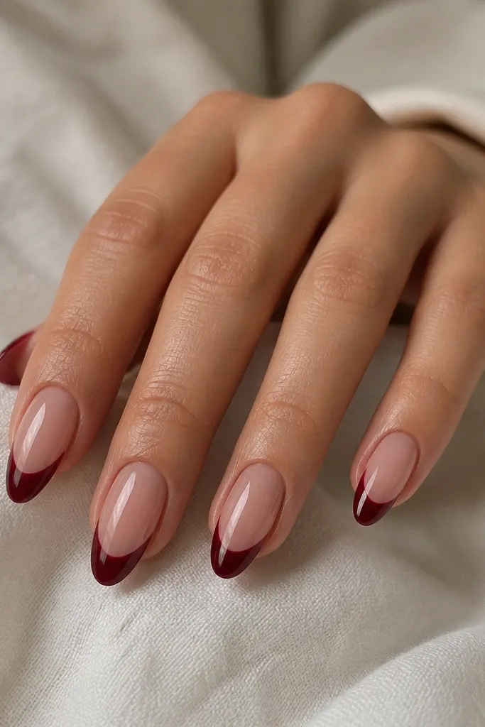 Almond nails with a sheer rosy nude base. The French tip is deep wine red but leaves a narrow negative-space crescent at the center of the smile line. The rest of the tip is glossy and sharply curved.