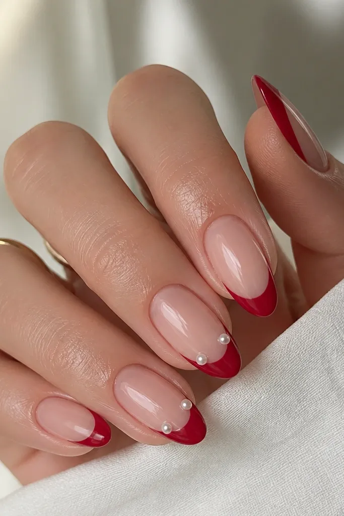 Almond nails with nude base and glossy red French tips. On two nails, there are tiny single pearl dots placed along the smile line - one dot on each side of the curve. The pearls look like small white beads.