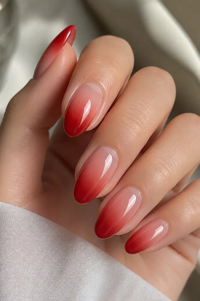 A set of medium soft-oval red extensions. The tips fade from deep cherry to a lighter red toward the middle, while the cuticle area stays clear with a neat half-moon shape. Everything is high-gloss and reflects light like glass.