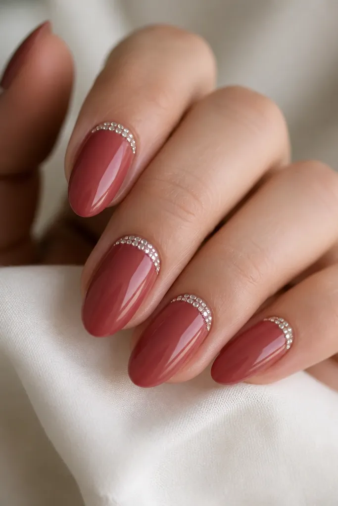 Medium almond nails with a glossy nude-red base. On each nail, a tight crescent of tiny clear rhinestones sits right at the cuticle line, leaving the rest smooth and glossy.