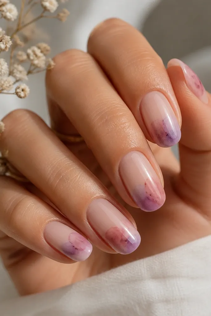 Nails with a nude base; a soft watercolor wash in dusty rose and light lilac sits near the tip and blends outward; a tiny darker splash dot sits in the center; glossy top coat.