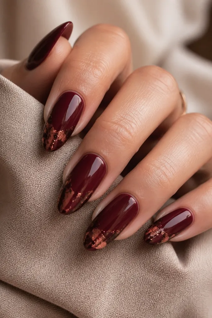 Almond nails with a solid wine red base and irregular foil at the tips in deep burgundy and copper tones; glossy top coat keeps the foil smooth.