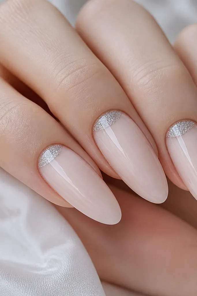 Close-up of long almond acrylic nails on fair skin. The base is sheer icy nude. The nail art is a thin band of fine silver micro-glitter only on the lower half-moon near the cuticle, with a clean bare nude line around it. Everything is glossy and smooth, no grainy texture.