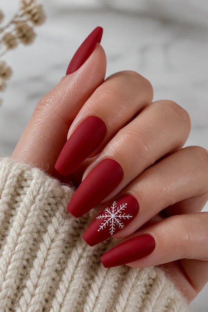 A set of medium coffin nails. Most nails are deep cherry red with a soft matte finish that looks like velvet. One accent nail has a white snowflake drawn with thin lines and tiny dot centers. The matte nails look smooth, with crisp edges near the cuticle.