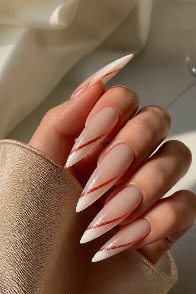 Long stiletto nails with a champagne nude to soft white ombre. A thin red candy stripe runs diagonally near the tip, and micro red shimmer surrounds it.