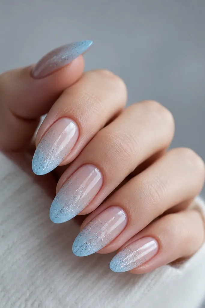 Long oval nails with an ice blue gradient fading into a clear, jelly-like center. Micro silver snow particles are scattered near the tip, all under a glossy top coat.