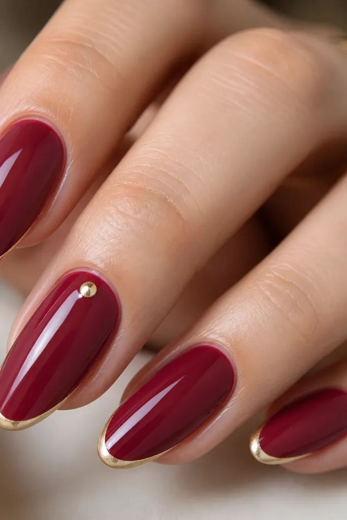 Close-up of almond nails painted in a glossy deep cranberry base. The tips have a thin, crisp French line in pale micro gold shimmer, with a clear glassy top coat that reflects light like jewelry. One nail shows a tiny gold dot at the cuticle area.