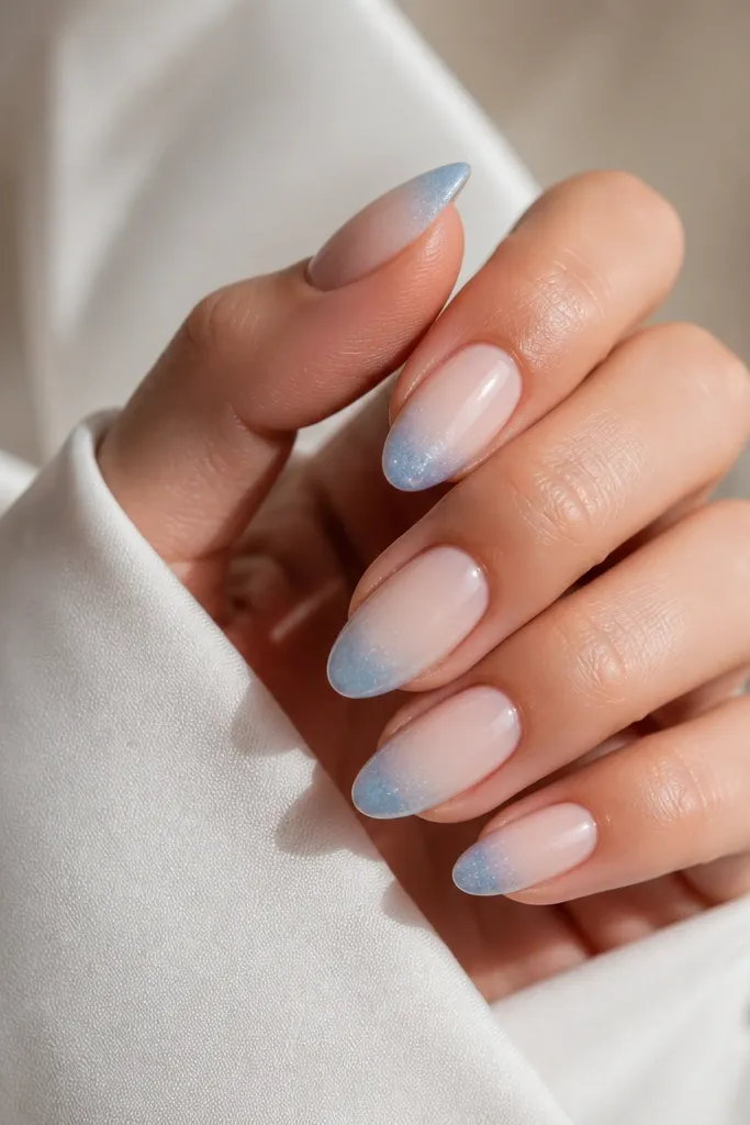 Medium almond nails with a sheer milky nude base. The tips fade into powder blue, starting thin near the cuticle and getting stronger at the edge. Over the whole tip area, there is a clear frost-like gel that makes tiny light sparkle.