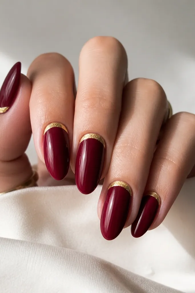 Almond nails with a deep cranberry red glossy base. On each nail, there are gold leaf half-moon shapes near the cuticle, like tiny crescents. The rest of the nail is clean and reflective.
