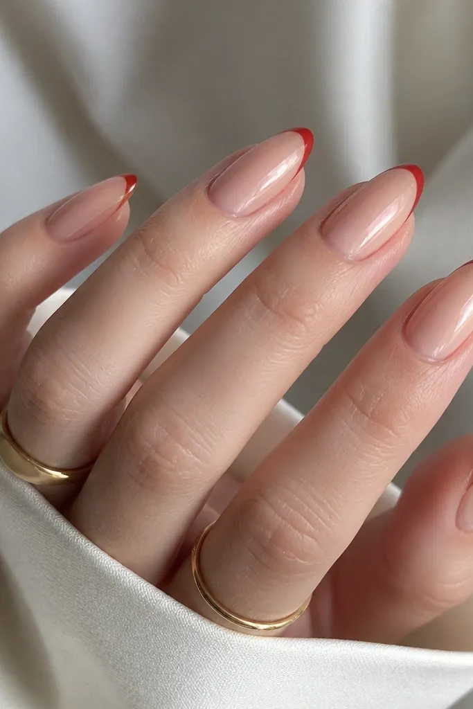 Almond nails on a fair skin hand. The base is a champagne nude glossy polish. A thin red French line runs along the tip edge, with no thick white border.