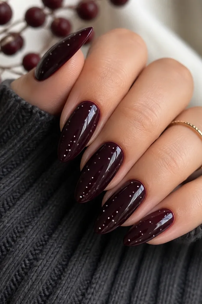 Almond nails in a deep black-cherry color with a glossy finish. Each nail has a few tiny white dots scattered like falling snow, clustered near the middle and tip.