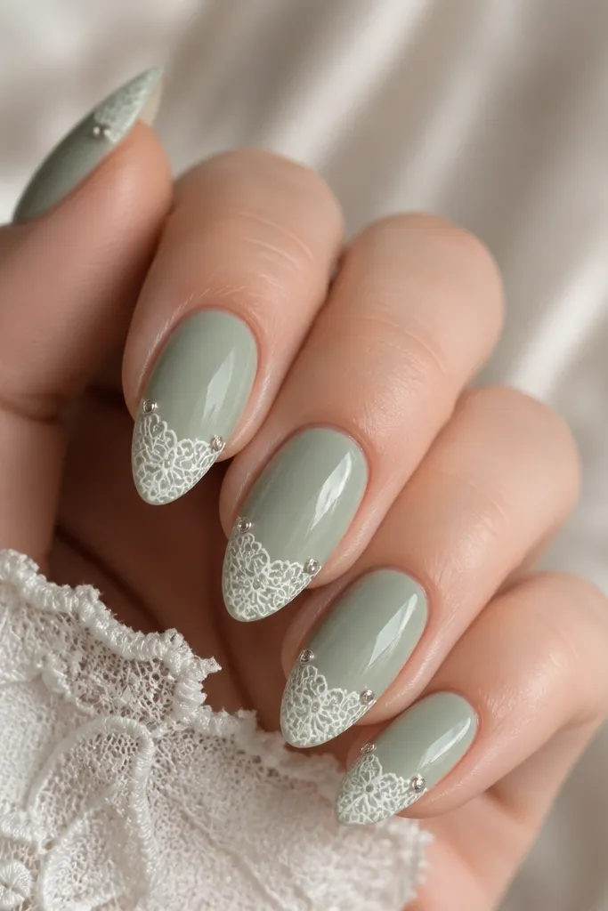 Almond nails with a soft sage-green base. The tips have delicate white lace patterning. One or two small clear rhinestones sit near the corner of the lace.