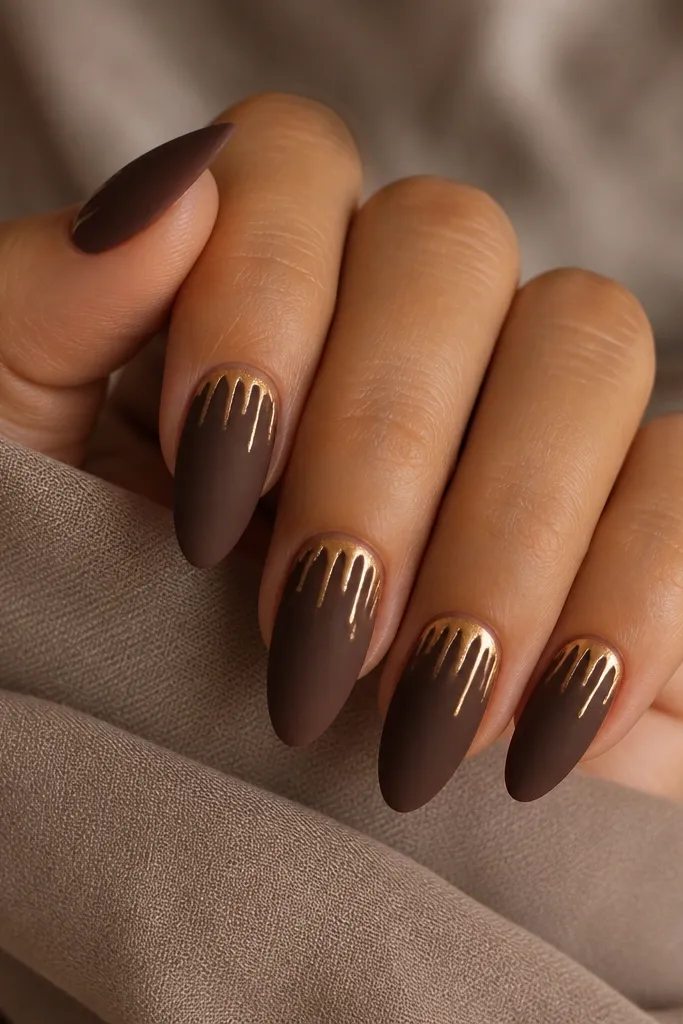 Almond nails with a matte cocoa-brown base. At the cuticle, there are thin gold drip lines that descend slightly down the nail, like melted gold icing.